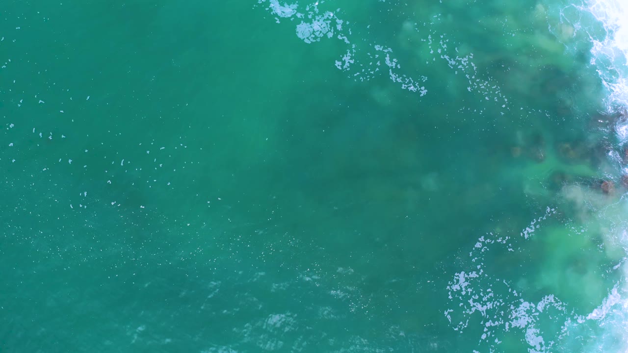Blue-green ocean surface in Los Cabos, Mexico, seen from above with scattered foam flecks and darker submerged patches, with no visible people.