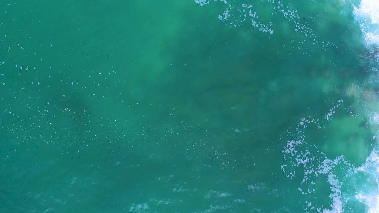 Teal water in Los Cabos, Mexico, with bright white breaking foam along the right edge and darker shapes under the surface, with no visible people.