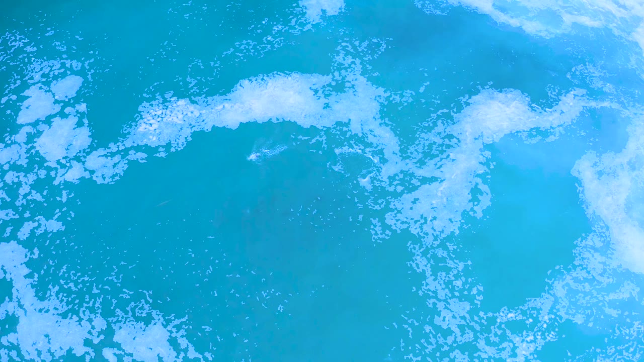 Overhead Los Cabos water frame with blue-green ocean filling the view, white foam breaking across the upper band, and no visible people.