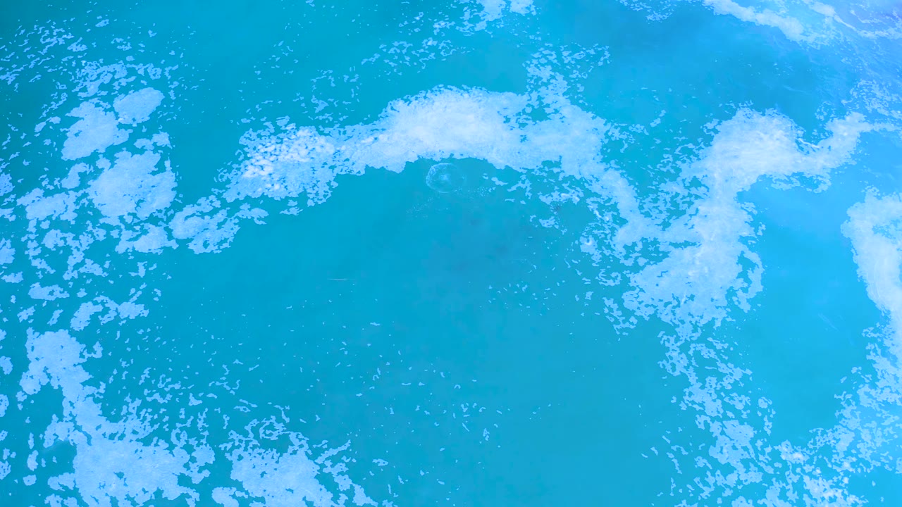 Overhead Los Cabos ocean frame with broken white foam crossing the center-left area over bright blue water and no visible people.