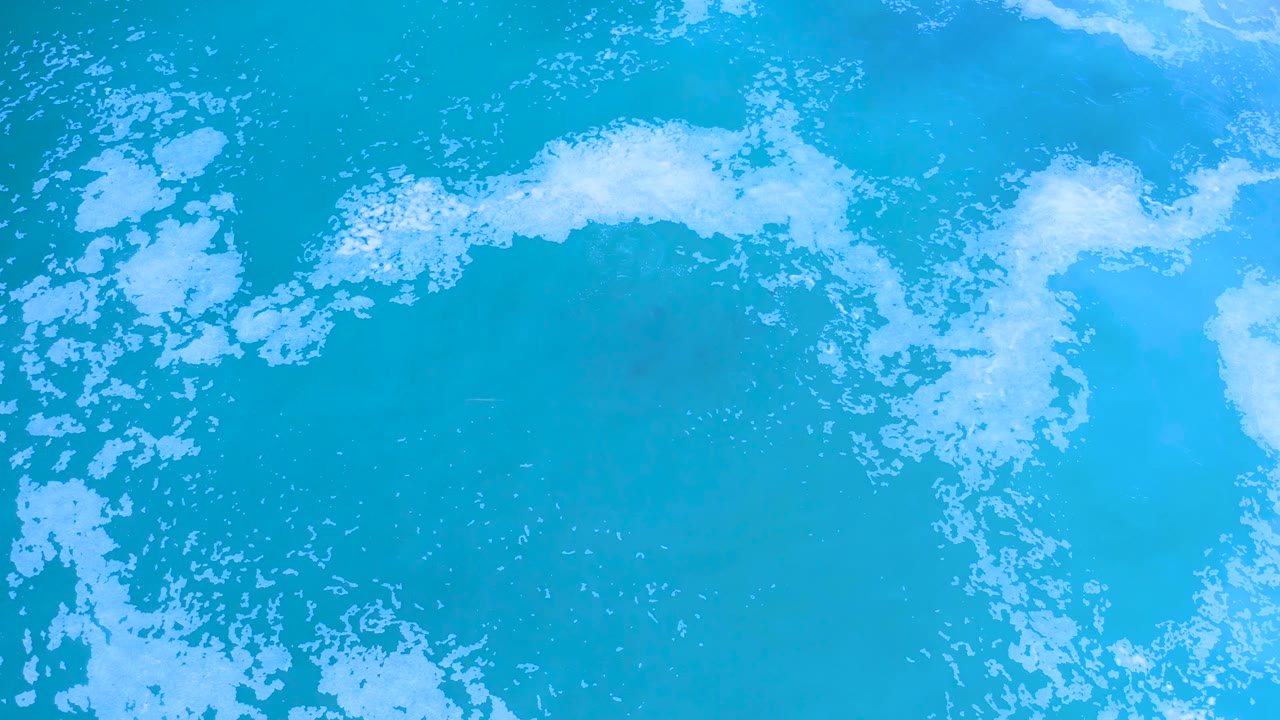 Overhead view of Los Cabos turquoise water with a curved foam streak near the upper center and a clearer blue area below, with no visible people.