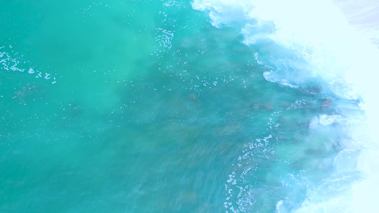 High overhead view of turquoise ocean in Los Cabos, where bright white surf covers the upper and right edges and darker patches show below the surface, with no visible people.