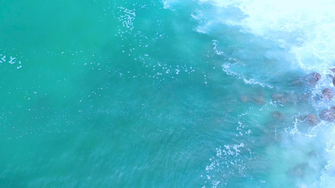 Aerial-looking frame of turquoise Los Cabos water with foam breaking around rocks along the right side, no visible people.