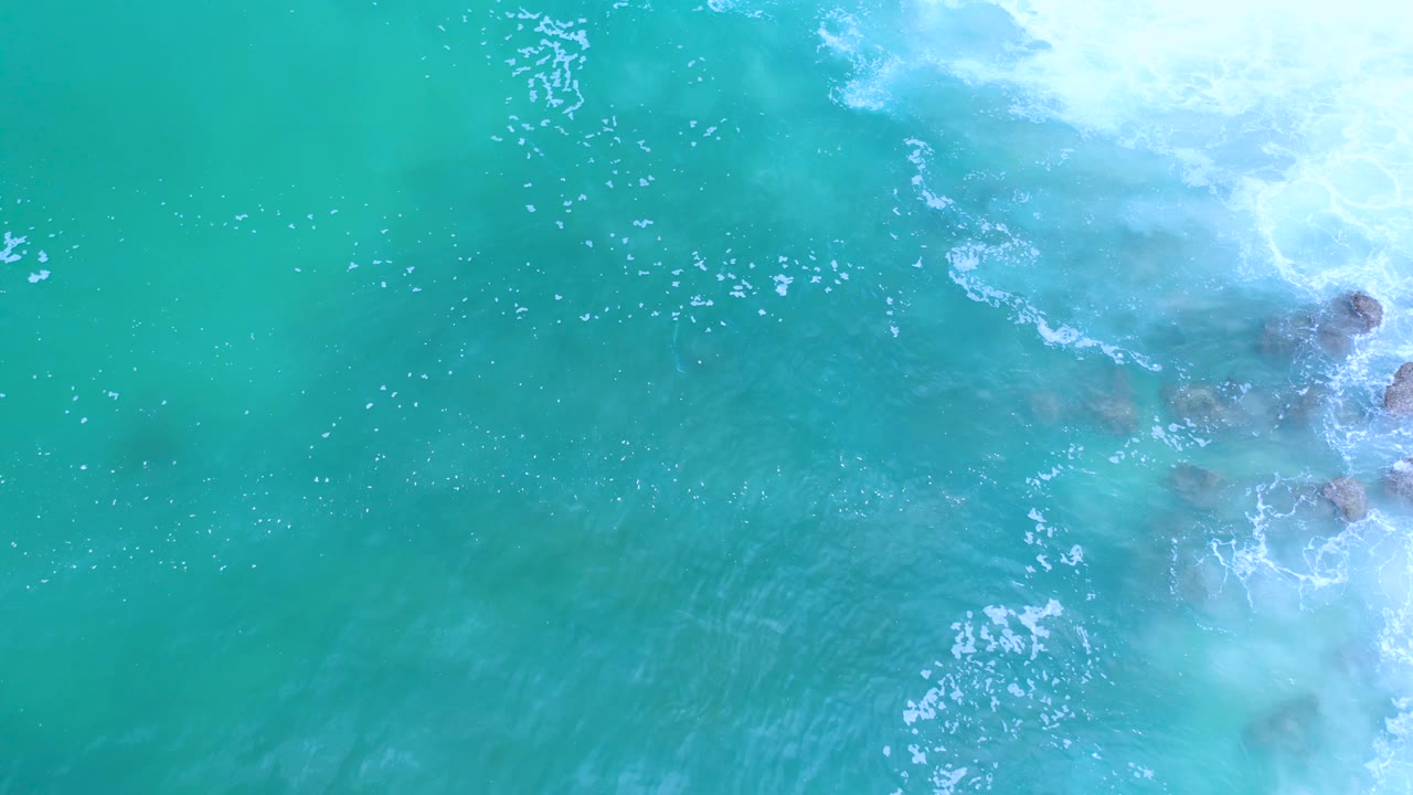 Overhead Los Cabos surf frame with pale foam at the upper right, blue-green water across the frame, and no visible people.