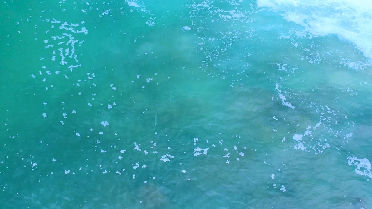 Overhead Los Cabos ocean frame showing translucent turquoise water, faint wave shadows, and foam collecting along the top edge with no visible people.