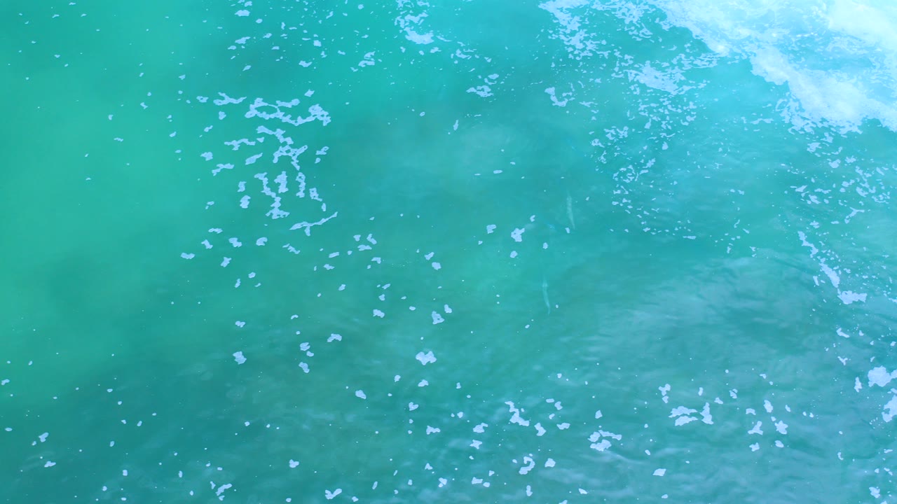 Overhead view of turquoise water in Los Cabos, Mexico, with scattered white foam near the upper right and no visible people.