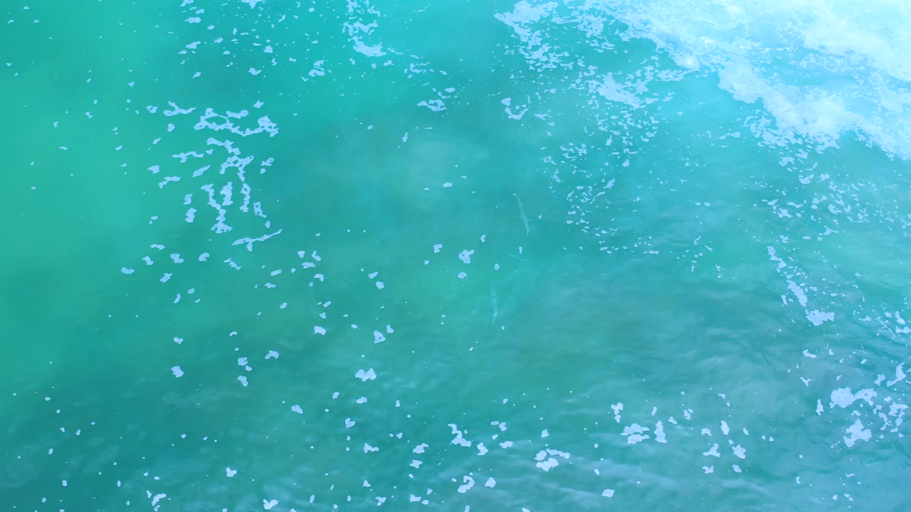 Aerial view of Los Cabos turquoise water, with brighter foam gathering along the top edge and no visible people.