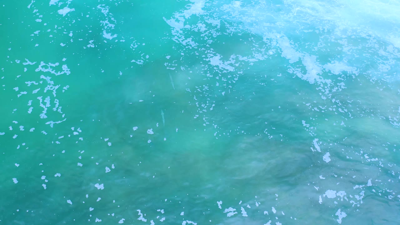 Aerial-style Los Cabos water frame with white foam clusters across the upper left, darker green patches below the surface, and no visible people.