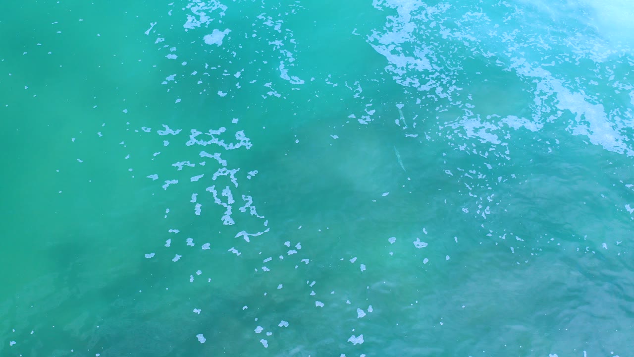 Overhead view of turquoise green ocean water in Los Cabos, Mexico, with scattered white bubbles near the upper right and no visible people.