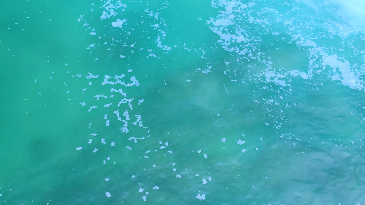 Overhead Los Cabos ocean frame with turquoise water, pale foam gathering along the upper right edge, and no visible people or boats.