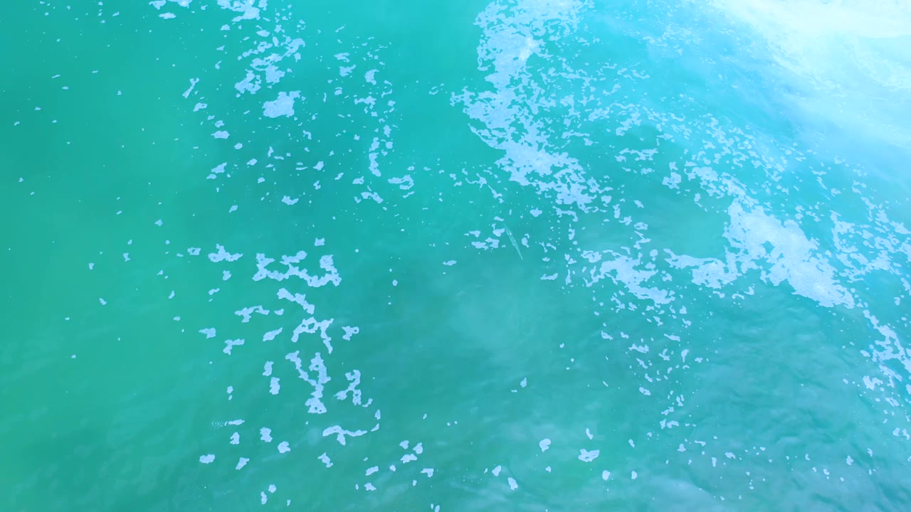 Overhead Los Cabos water frame with a loose streak of white bubbles running through blue-green ocean and no visible people.