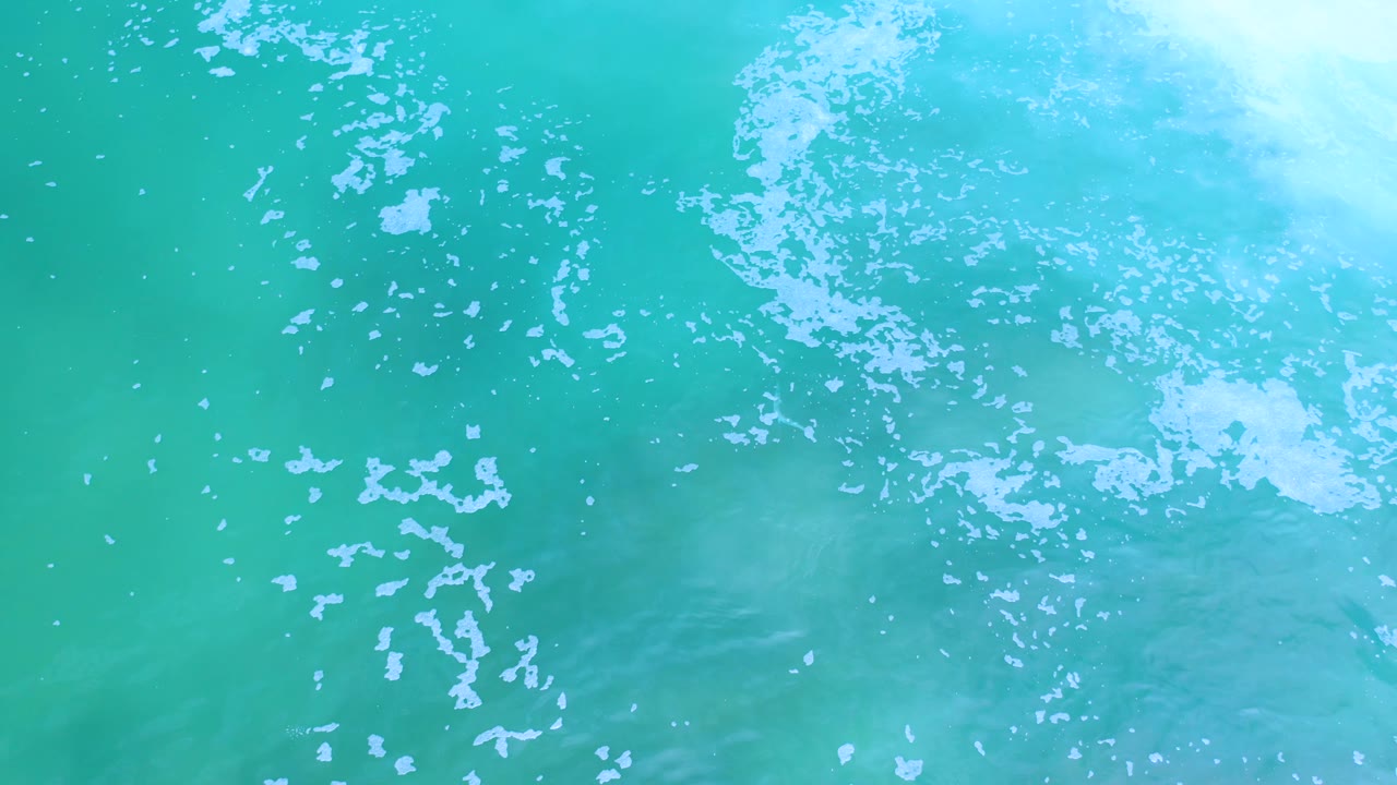Close overhead view of Los Cabos turquoise ocean, with pale white foam clustered near the upper edge and no visible people.