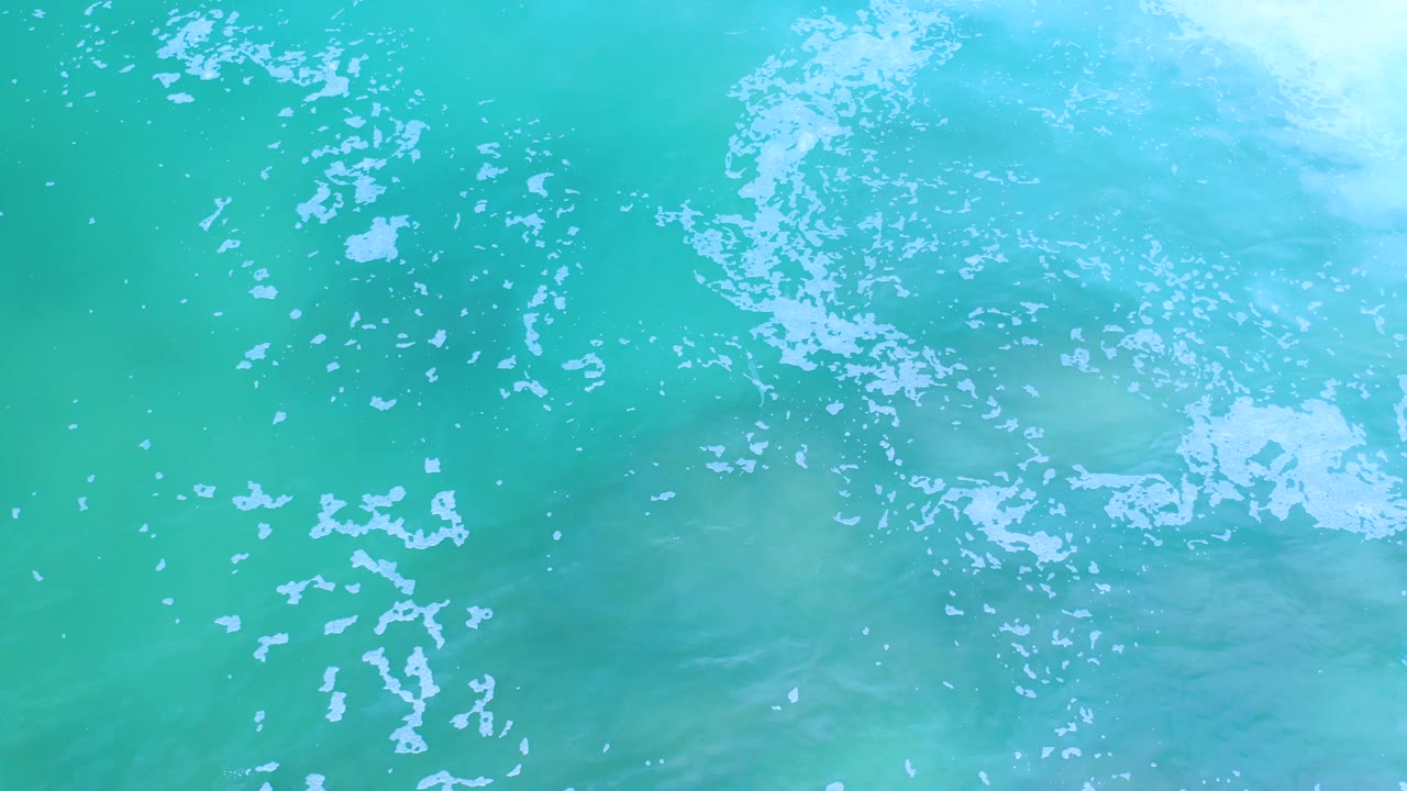 Overhead Los Cabos ocean detail with white bubble clusters spread across blue-green water and no visible people.
