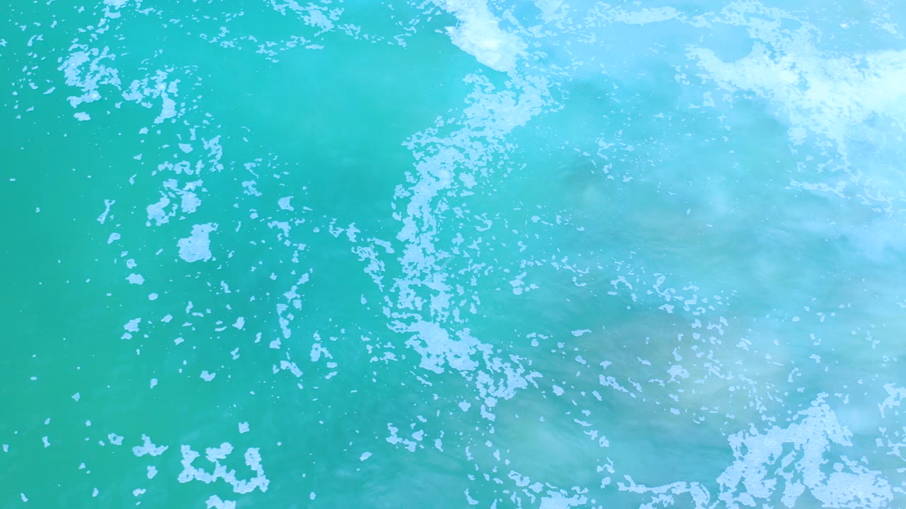 Overhead view of turquoise ocean water in Los Cabos, Mexico, with scattered white foam across the upper right and no visible people.