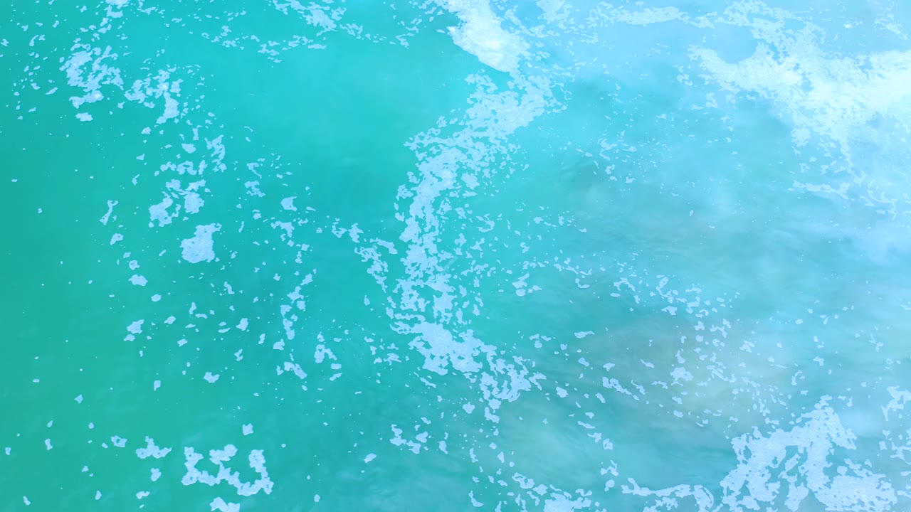 Aerial view of blue-green Los Cabos ocean water with pale foam streaks drifting across the top of frame and no visible people.