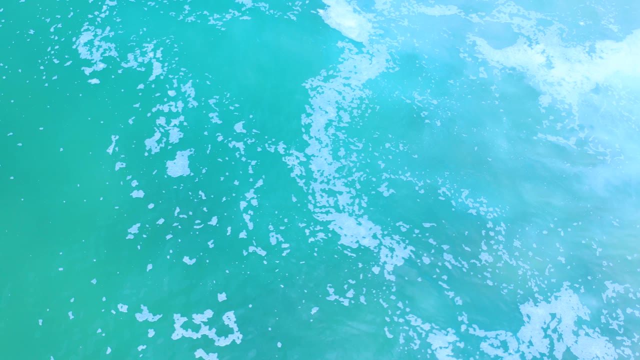 Top-down Los Cabos ocean frame with aqua water, scattered white foam clusters, and no visible people, boats, or shoreline.