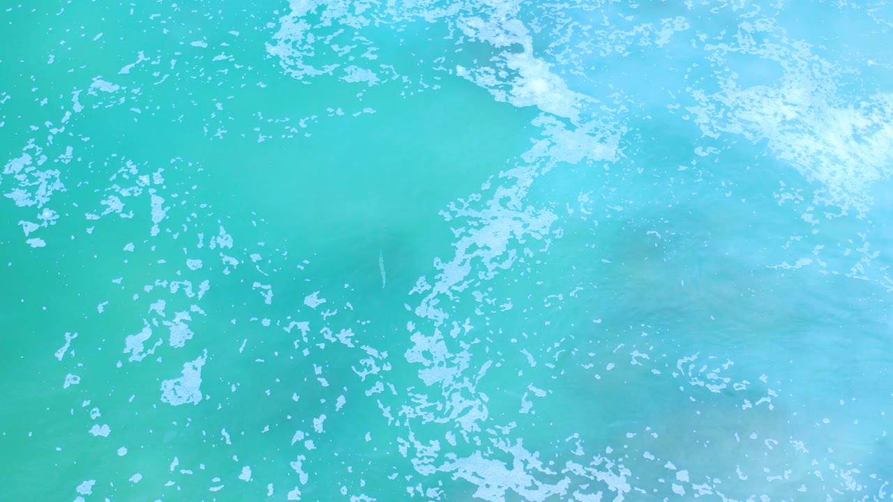 Overhead view of turquoise shallow ocean water in Los Cabos, Mexico, with white foam spread across the upper edge and no visible people.
