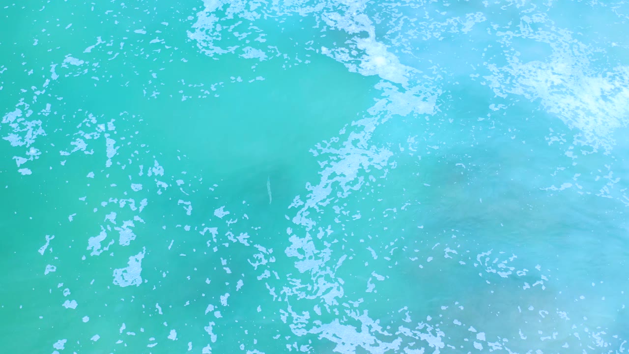 White foam drifts across clear turquoise water in Los Cabos, Mexico, with a pale rocky seabed visible beneath and no visible people.