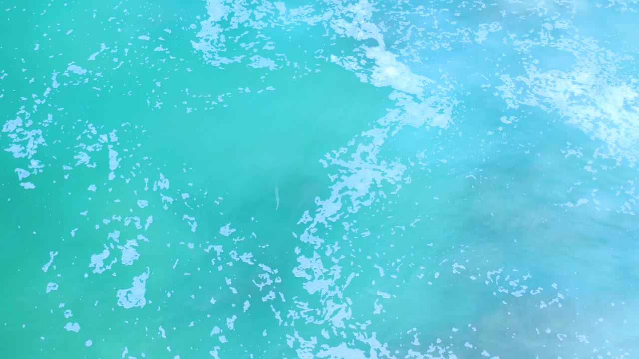 A brighter patch of white sea foam sits near the upper right of shallow green-blue water in Los Cabos, Mexico, with no visible people.