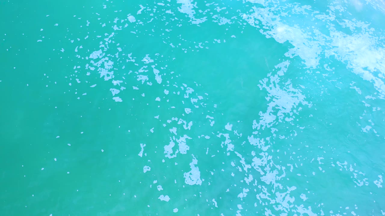 Overhead Los Cabos sea surface with aqua and green water tones, white foam spread diagonally across the top right, and no visible people.
