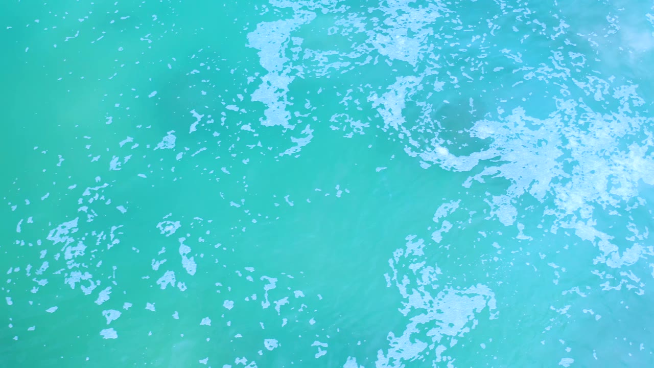Overhead Los Cabos water frame with broken white foam spread across the upper half and clear turquoise water below, with no visible people.
