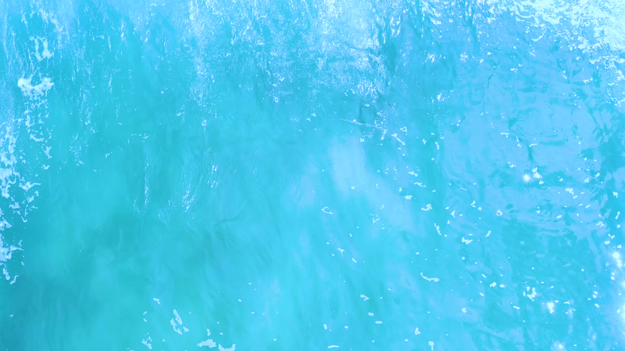 A top-down frame of blue-green Los Cabos sea shows a narrow vertical streak of white foam crossing the water, with no visible people.
