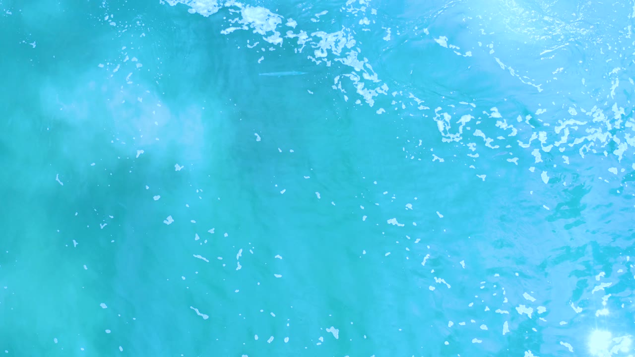 A top-down view of turquoise Los Cabos water shows diffuse light rays, cloudy blue-green texture, white surface speckles, and no visible people.