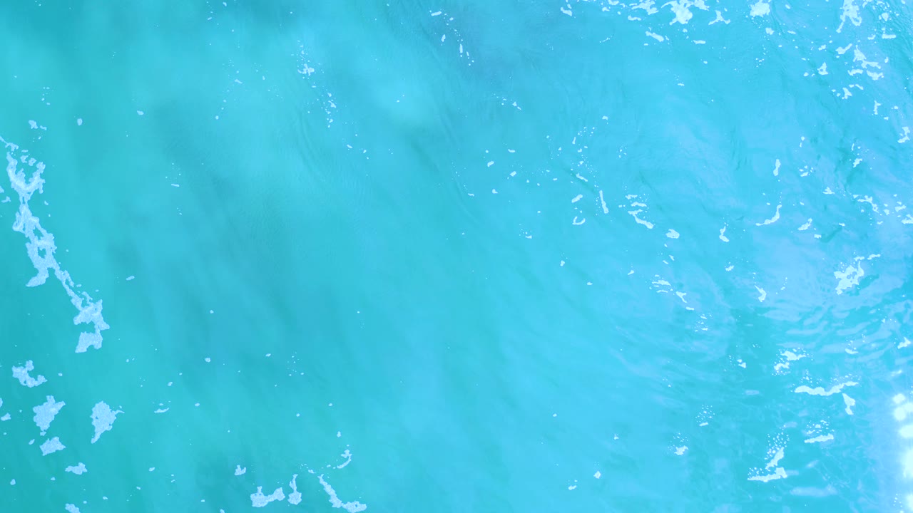 Overhead view of blue water in Los Cabos, Mexico, where bright sun shimmer and thin vertical foam streaks stand out, with no visible people.