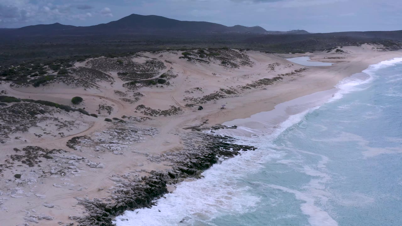 Drone-height view of Los Cabos surf pushing along the rocky shore, with pale beach and dune ridges leading back to low scrub and a dark mountain range, and no visible people.