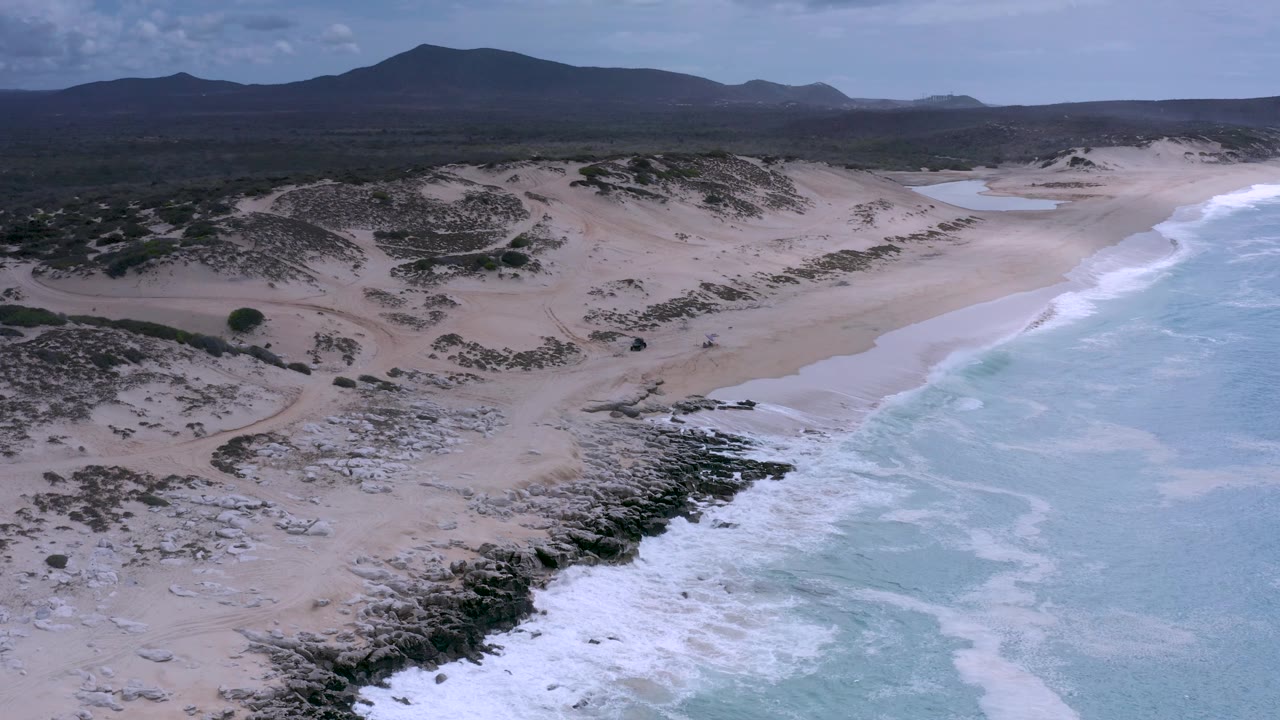 Wide Los Cabos aerial frame with blue-green water covering the right side, white foam curling into shore, sandy dunes in the middle distance, mountains beyond, and no visible people.
