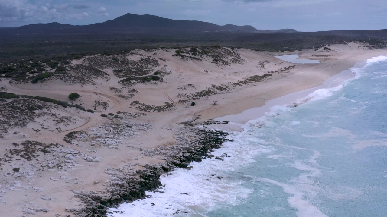 Los Cabos coastline from above with dark rocks in the lower frame, surf breaking across the right edge, open sand and dunes beyond, distant mountains, and no visible people.