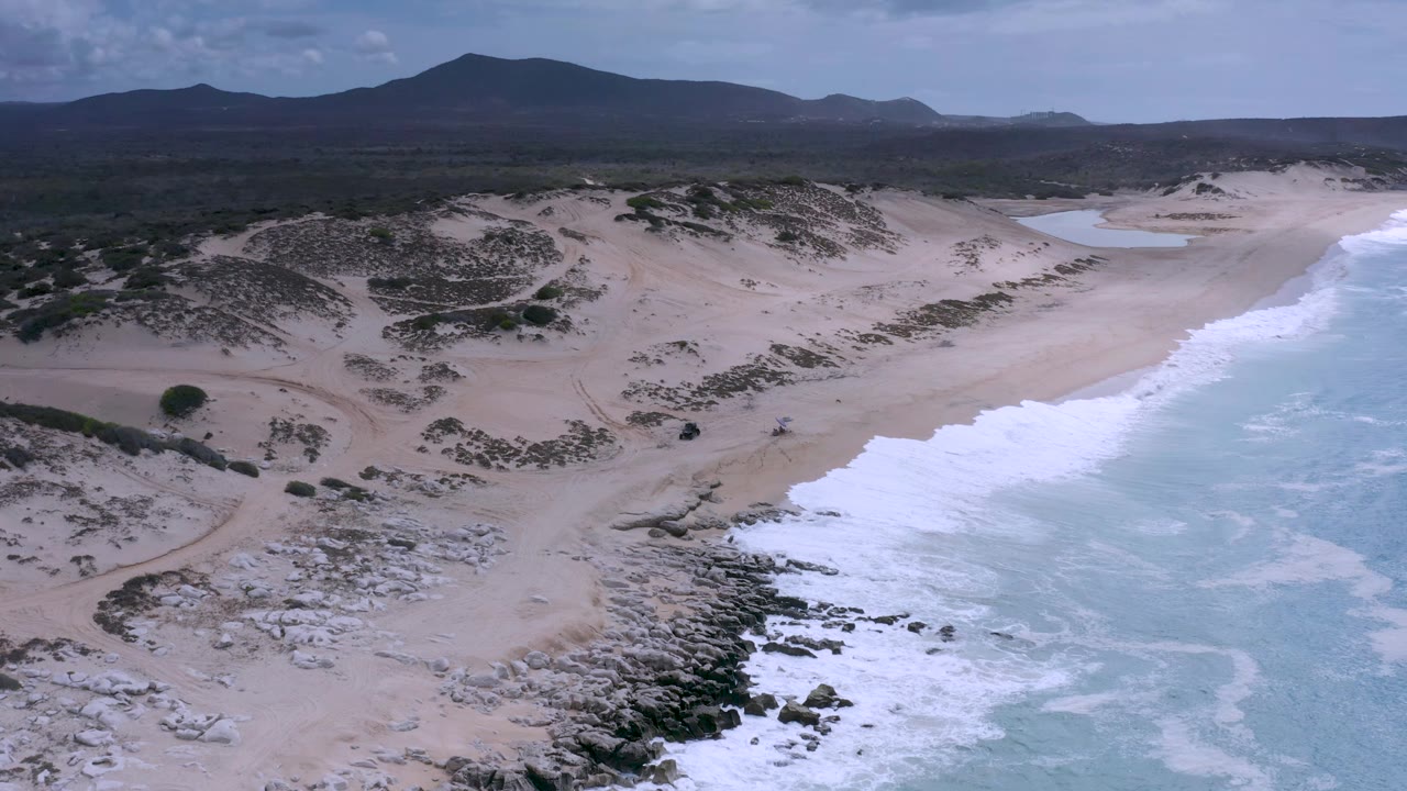 Aerial frame from Los Cabos, Mexico, with a bright band of white surf curving up the beach, dark rocks at the lower edge, dune tracks inland, mountain silhouettes, and no visible people.