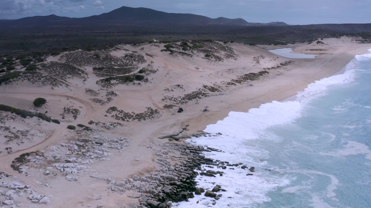 Los Cabos, Mexico aerial view with foamy ocean filling the right side, pale dunes and beach through the center, a small inland water patch near the top, distant dark hills, and no visible people.