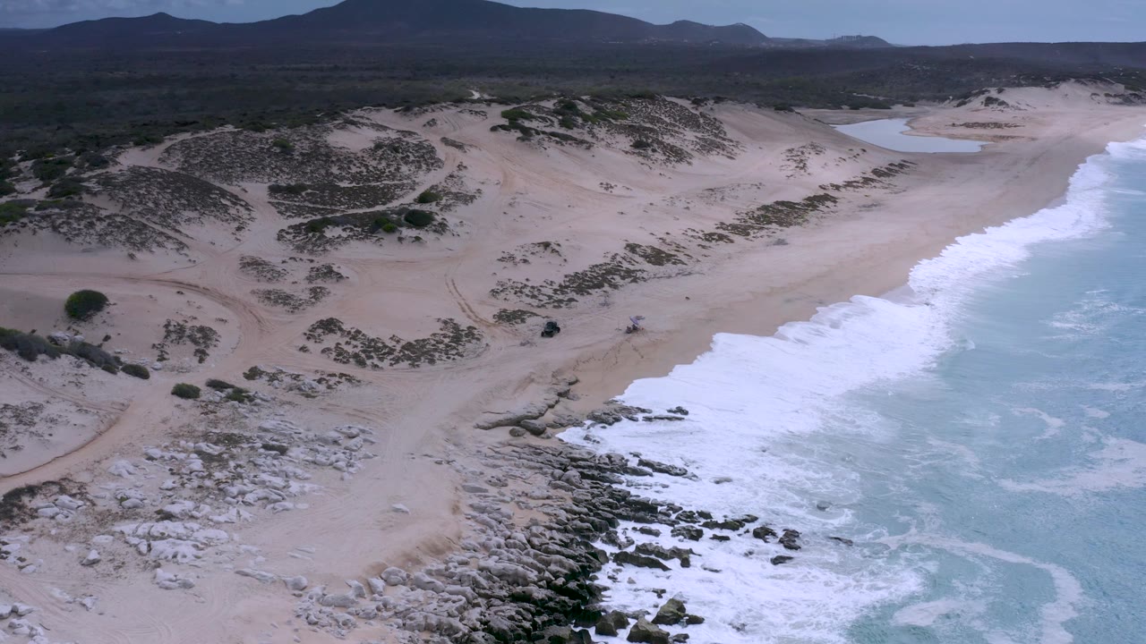 Drone frame over Los Cabos, Mexico, with surf washing onto the right edge of a pale sandy beach, darker dune scrub across the middle, rocky outcrops below, mountains in the distance, and no visible people.