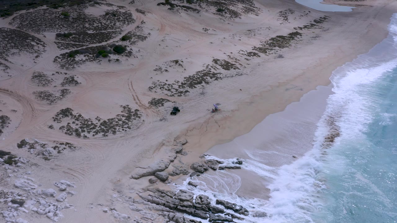 Aerial Los Cabos shoreline frame with foamy water running along the right side, rocks at the lower beach, curved tire tracks in the sand, dune scrub above, and no visible people.