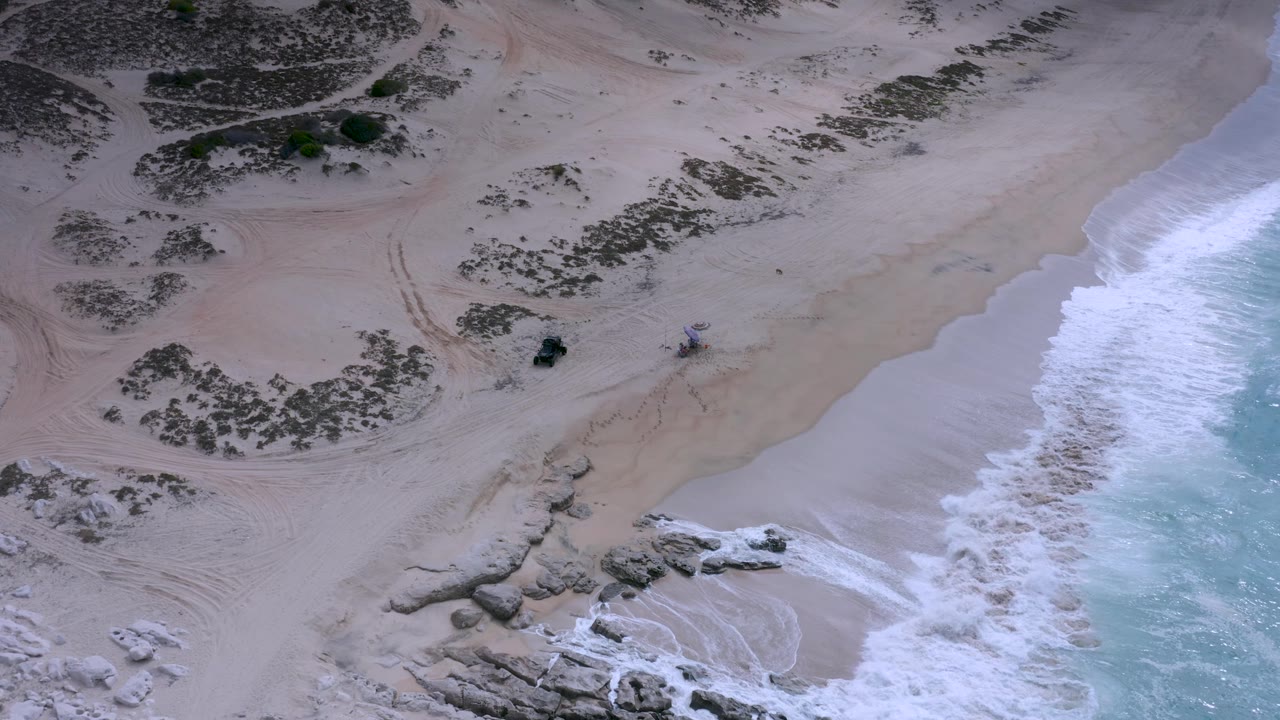 Overhead view of the Los Cabos beach with the surf line pushed farther right, open sand filling the middle, scrub-covered dunes at top left, rock clusters below, and no visible people.