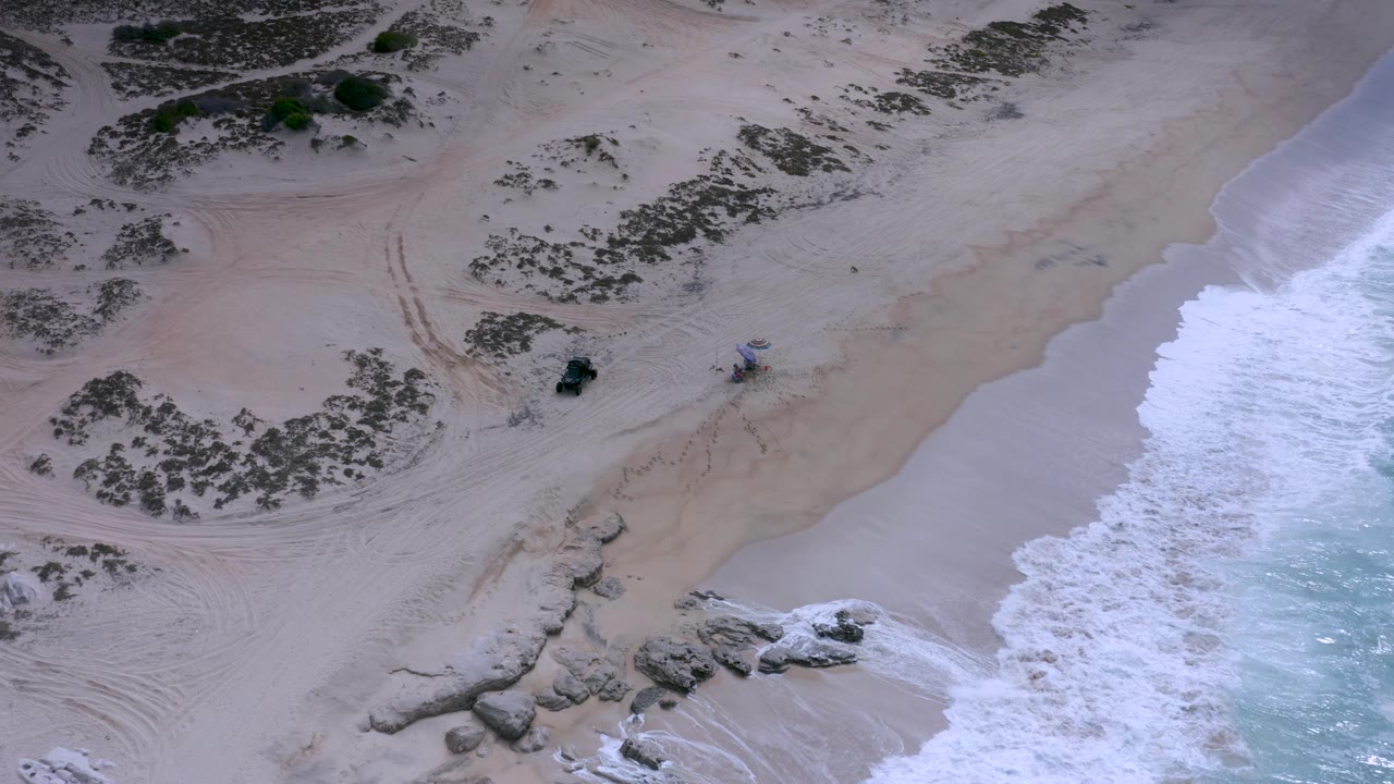 Los Cabos aerial beach view with a broad band of white wave wash on the right, textured sand and tire marks across the center, darker vegetation patches on the left, and no visible people.