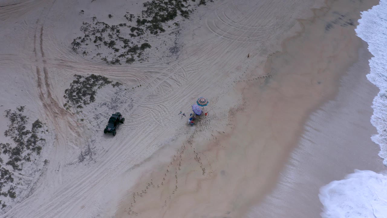 Top-down aerial frame over a Los Cabos beach shows pale sand, surf at the right edge, a dark Jeep-like vehicle, colorful umbrellas, and tiny people near the setup.