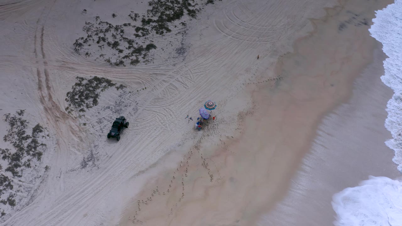 Aerial beach frame with umbrellas, small people, surf, sand tracks, and parked vehicle.