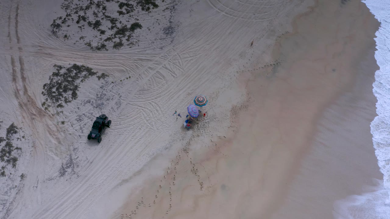 Los Cabos aerial frame shows a dark Jeep-like vehicle on tracked sand, a small umbrella cluster with people nearby, scrub patches at the top and left, and white waves along the right side.
