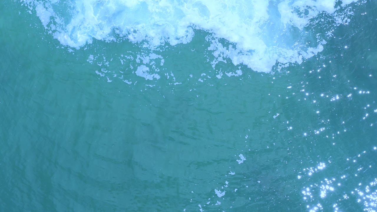Overhead view of turquoise water in Los Cabos, Mexico, with white foam near the top edge, scattered sun sparkles on ripples, and no visible people.