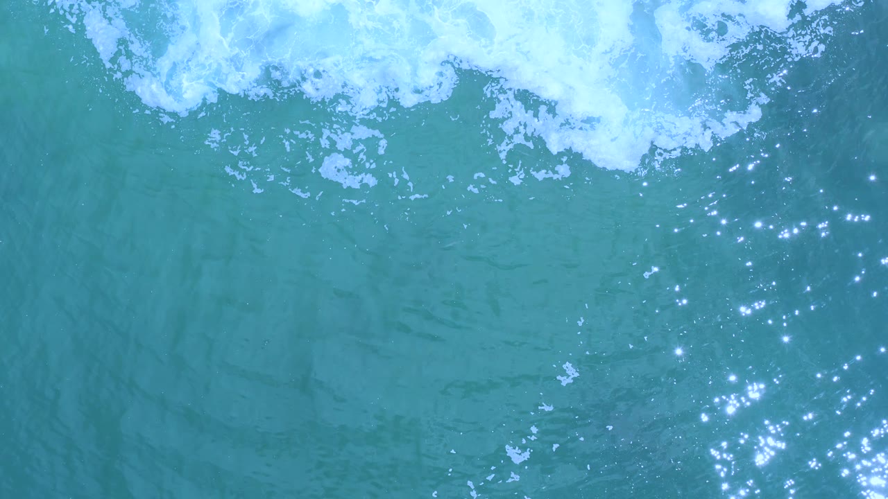 Overhead Los Cabos ocean view with a broad white foam band across the top, blue-green water below, fewer bright glints, and no visible people.