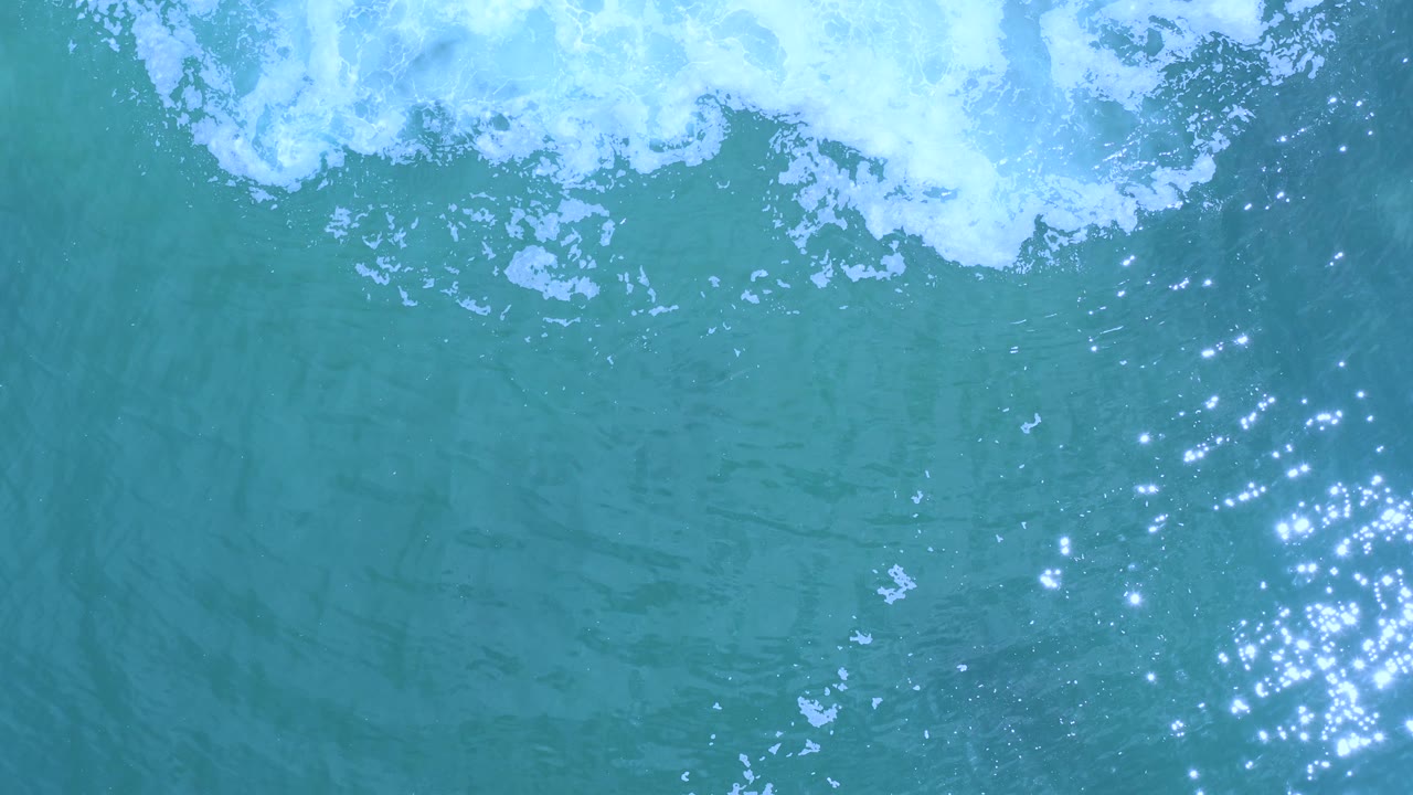 Overhead frame from Los Cabos, Mexico, showing turquoise water, white foam along the upper edge, dense sun sparkle clusters at right, and no visible people.