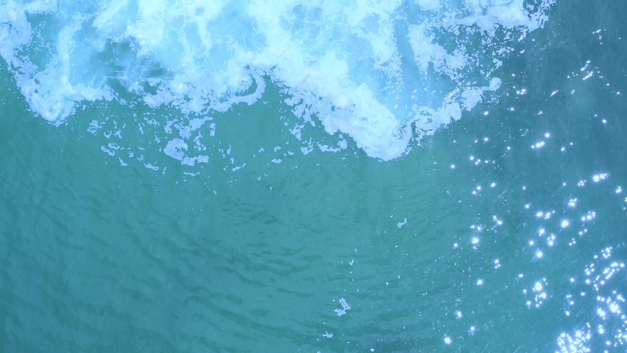 Top-down blue ocean with white surf and sun glints.