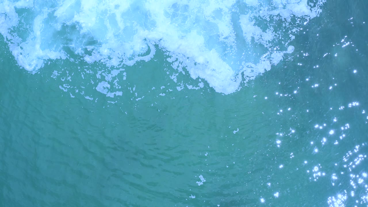Top-down Los Cabos ocean frame with a broad white foam band near the top, teal water beneath it, small surface ripples, and no visible people.
