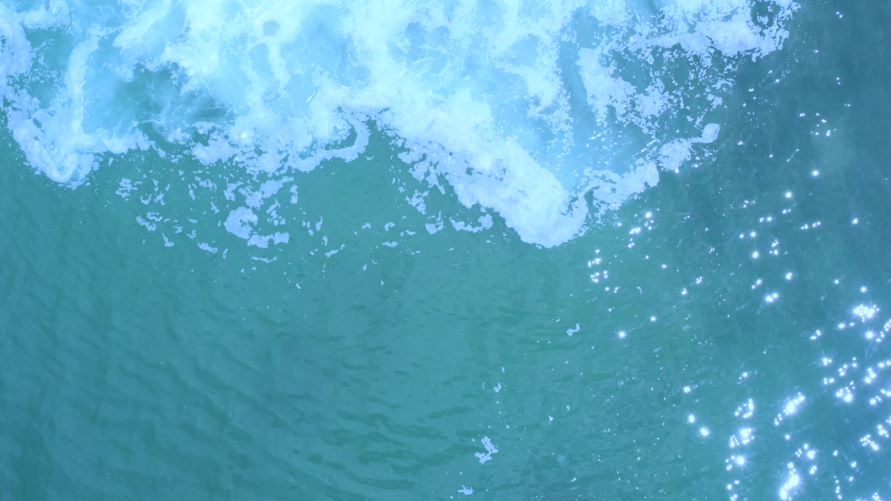 Overhead blue sea in Los Cabos, Mexico, where broken white surf sits along the upper edge and clusters of bright light reflect across the right side, with no visible people.
