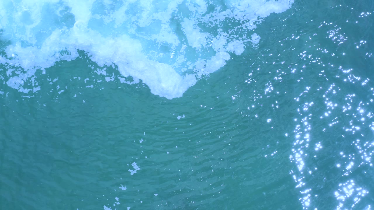 Overhead Los Cabos ocean frame with a white foam band at the top and a dense cluster of sunlight reflections crossing blue-green ripples, no visible people.