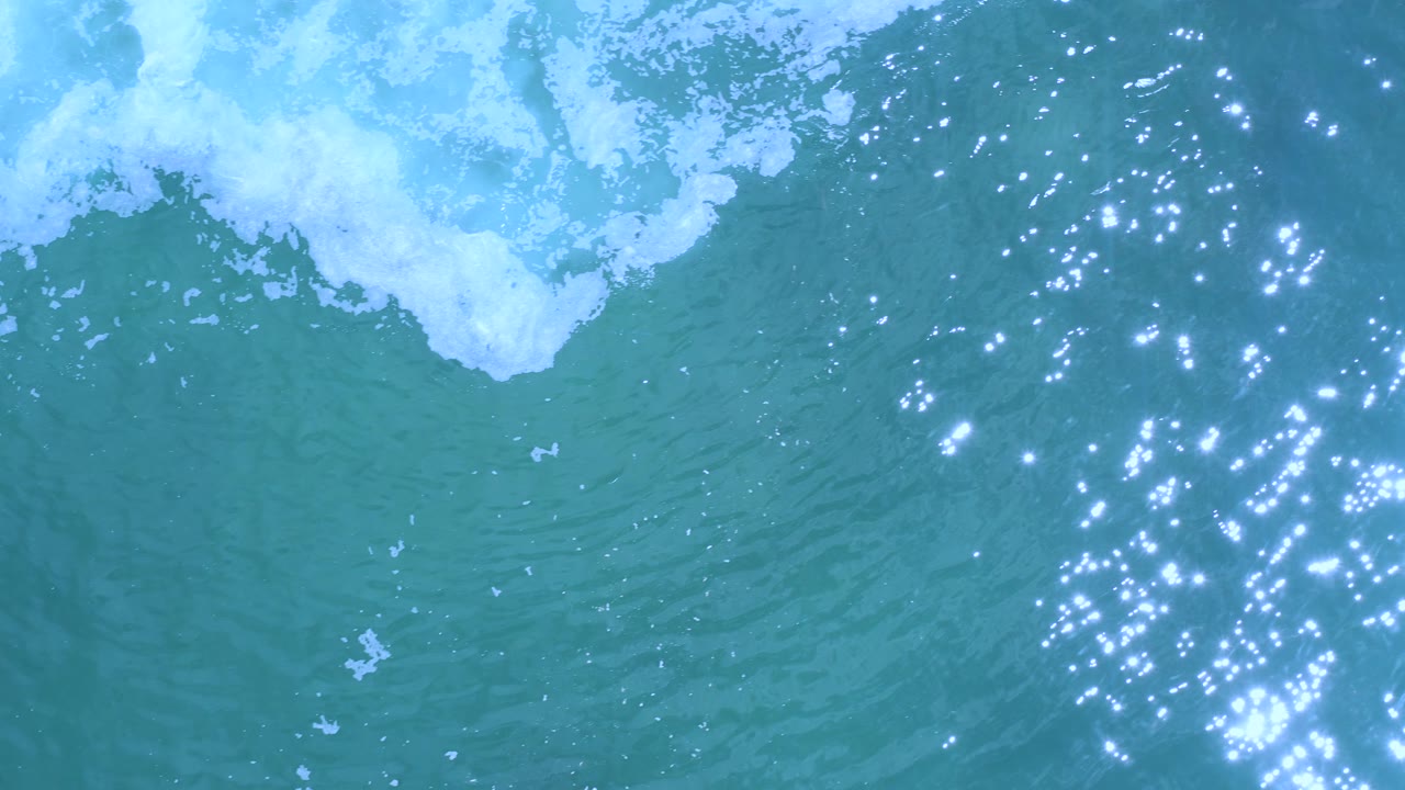 Aerial view of small wave foam pushing across turquoise water in Los Cabos, Mexico, with scattered bright reflections on the right side, no visible people.