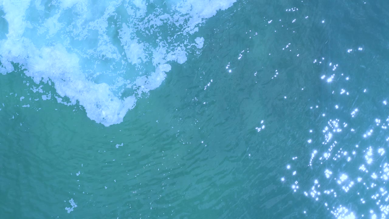 Overhead Los Cabos water detail with foam crossing the upper frame, green-blue ocean below, and a bright cluster of sun glints near the right edge, no visible people.