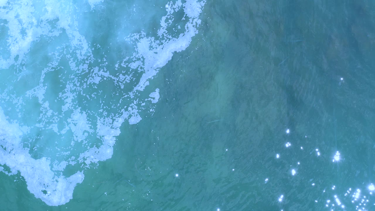 Overhead view of turquoise seawater in Los Cabos, Mexico, with white foam near the upper left and scattered sun reflections, no visible people.